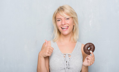 Caucasian adult woman over grey grunge wall eating chocolate donut happy with big smile doing ok sign, thumb up with fingers, excellent sign