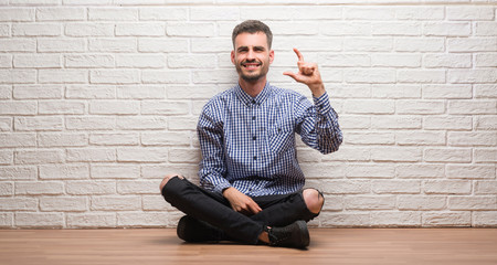 Young adult man sitting over white brick wall smiling and confident gesturing with hand doing size sign with fingers while looking and the camera. Measure concept.