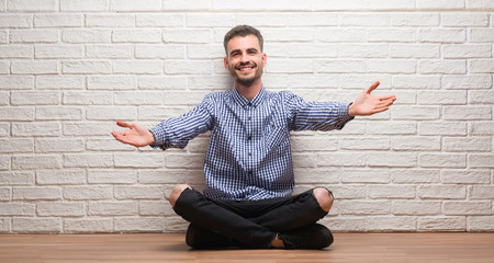 Young adult man sitting over white brick wall looking at the camera smiling with open arms for hug. Cheerful expression embracing happiness.