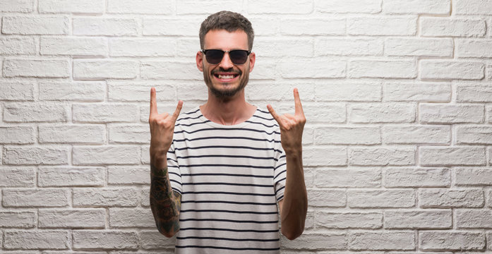 Young adult man wearing sunglasses standing over white brick wall shouting with crazy expression doing rock symbol with hands up. Music star. Heavy concept.