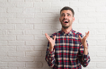 Young adult man standing over white brick wall crazy and mad shouting and yelling with aggressive expression and arms raised. Frustration concept.