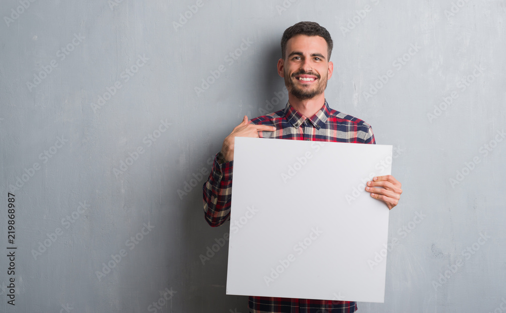 Wall mural young adult man over brick wall holding banner with surprise face pointing finger to himself