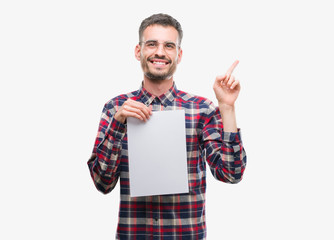 Young hipster adult man holding blank paper sheet very happy pointing with hand and finger to the side