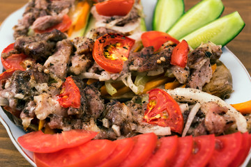 Fried meat with tomatoes, onions and carrots, laid on baked eggplant in a plate next to sliced fresh cucumbers and tomatoes, close-up.