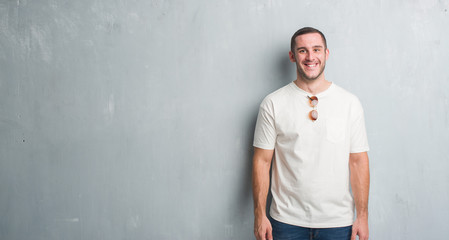 Young caucasian man over grey grunge wall wearing sunglasses with a happy and cool smile on face. Lucky person.