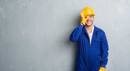 Young caucasian man over grey grunge wall wearing contractor uniform and safety helmet with happy face smiling doing ok sign with hand on eye looking through fingers