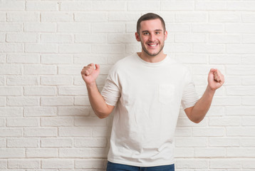 Young caucasian man standing over white brick wall celebrating surprised and amazed for success with arms raised and open eyes. Winner concept.