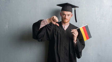 Young redhead man over grey grunge wall wearing graduate uniform holding germany flag with angry face, negative sign showing dislike with thumbs down, rejection concept