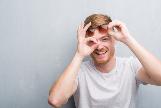 Young redhead man over grey grunge wall wearing retro sunglasses with happy face smiling doing ok sign with hand on eye looking through fingers