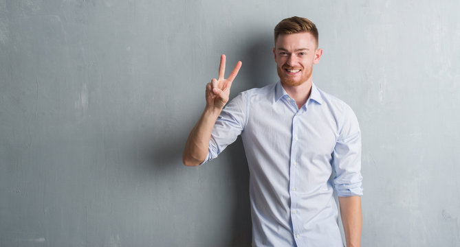 Young redhead business man over grey grunge wall showing and pointing up with fingers number two while smiling confident and happy.