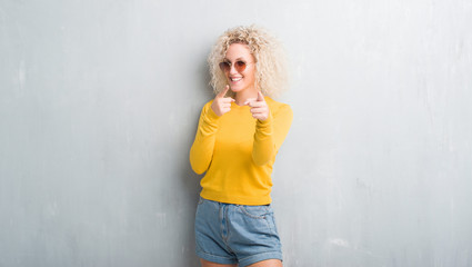 Fototapeta premium Young blonde woman with curly hair over grunge grey background pointing fingers to camera with happy and funny face. Good energy and vibes.