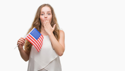 Young blonde woman holding flag of USA cover mouth with hand shocked with shame for mistake, expression of fear, scared in silence, secret concept