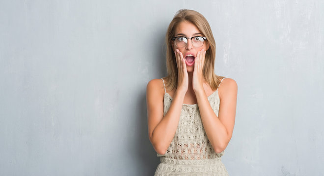 Beautiful young woman standing over grunge grey wall wearing glasses scared in shock with a surprise face, afraid and excited with fear expression