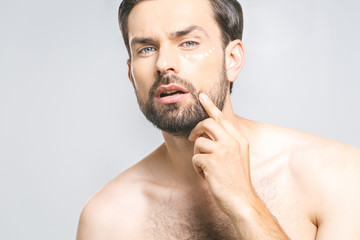 Obraz premium Skin care. Handsome happy young shirtless man applying cream at his face and looking up while standing over gray background and looking at camera. Close-Up. Space for text.