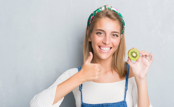 Beautiful young woman over grunge grey wall eating fresh kiwi happy with big smile doing ok sign, thumb up with fingers, excellent sign
