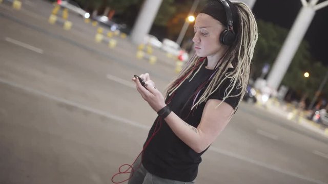 Young attractive woman with dreadlocks use smartphone listening music on headphones. On street at night 