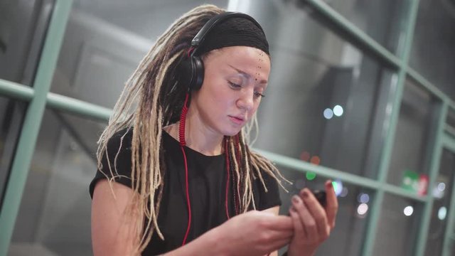 Young attractive woman with dreadlocks use smartphone listening music on headphones. On street at night 