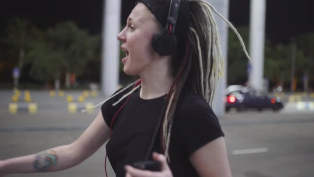 Young attractive woman with dreadlocks use smartphone listening music on headphones. On street at night 