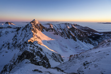 High winter alpine like mountain landscape covered with snow. Sunrise sky, yellow, orange, purple and blue colours. Rocky mountain ridge and high peaks of High Tatras, Slovakia.