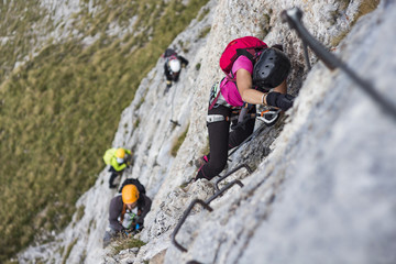 Woman and group of other people climbing on steep rock face on via ferrata. Climbers on via ferrata climbing route. Alpine ferrata ascent to summit. Summer adventure mountain activity.