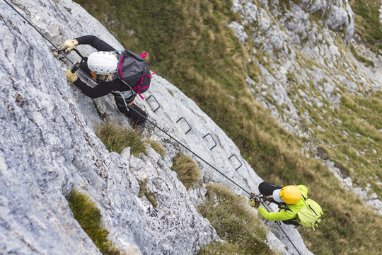 Woman and group of other people climbing on steep rock face on via ferrata. Climbers on via ferrata climbing route. Alpine ferrata ascent to summit. Summer adventure mountain activity.