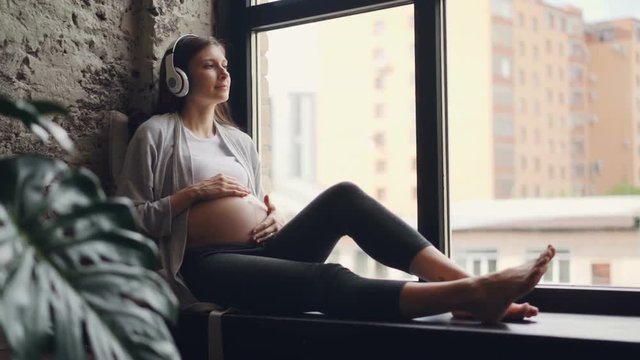 Beautiful girl expecting mother is caressing her tummy and listening to music wearing wireless headphones sitting on window sill in apartment. Love, family and technology concept.