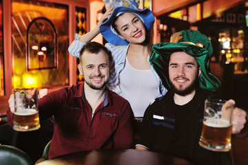 friends - boys and girls in Bavarian hats drinking beer on the background of the bar during the celebration of Oktoberfest or St. Patrick's Day
