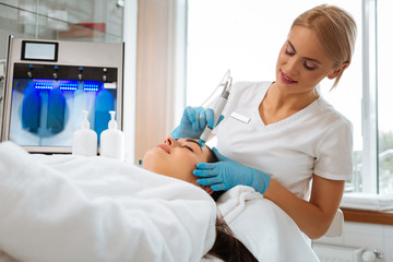 My workplace. Delighted nice cosmetologist standing near her client while doing a beauty procedure