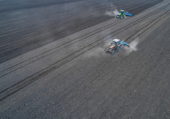 Aerial view of two blue tractors plows the earth in field on a summer day against a black earth...