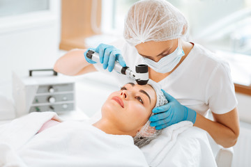 Newest equipment. Nice skilled dermatologist holding a dermatoscope while working with her patient