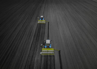 Aerial view of two blue tractors plows the earth in field on a summer day against a black earth...