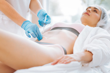 Body treatment. Skilled professional beautician holding a syringe while doing a body injection