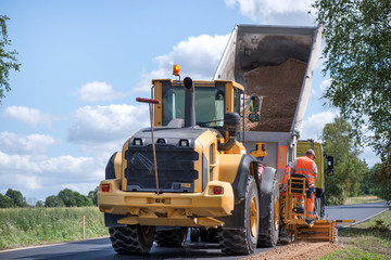 Road construction workers repairing highway road on sunny summer day. Loaders and trucks on newly made asphalt. Heavy machinery working on street. Road curbs being constructed with gravel