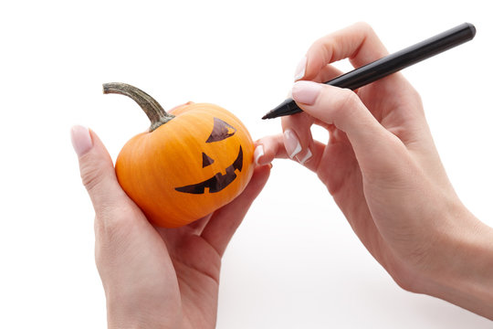 Woman's hands drawing scary face on little pumpkin with a black marker pen isolated on white background for Halloween. Holiday decoration concept.