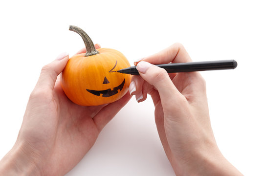 Woman's hands drawing scary face on little pumpkin with a black marker pen isolated on white background for Halloween. Holiday decoration concept.