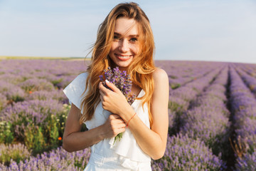 Photo of happy young woman in dress holding bouquet with flowers, while walking outdoor through lavender field in summer