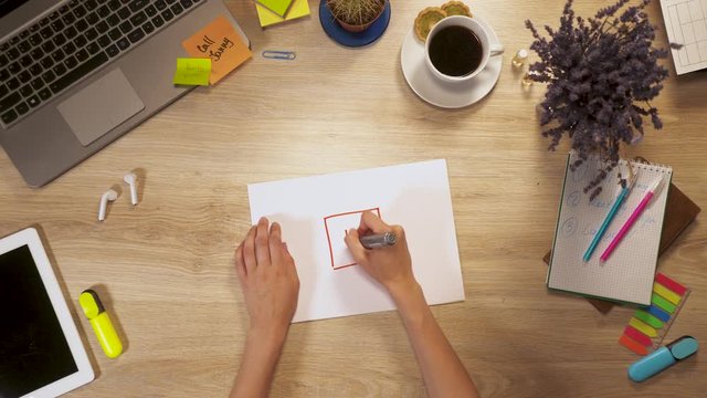 Girl draws a house on a sheet of paper, sitting at work table, first-person view of hands.