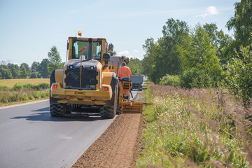 Road construction workers repairing highway road on sunny summer day. Loaders and trucks on newly made asphalt. Heavy machinery working on street. Road curbs being constructed with gravel
