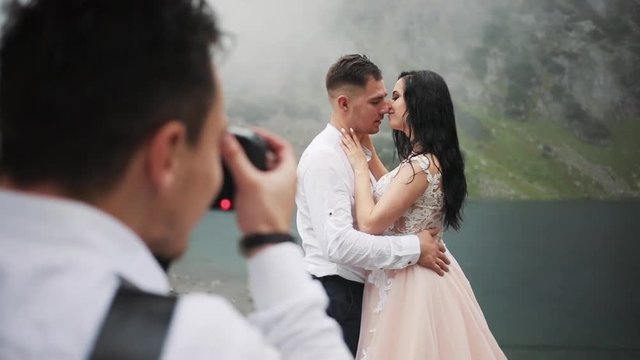 Close up photographer taking pictures lovely wedding couple stands near lake in mountains, hugging kissing on a cloudy day groom happy portrait dress celebration party face bridge young nature outdoor