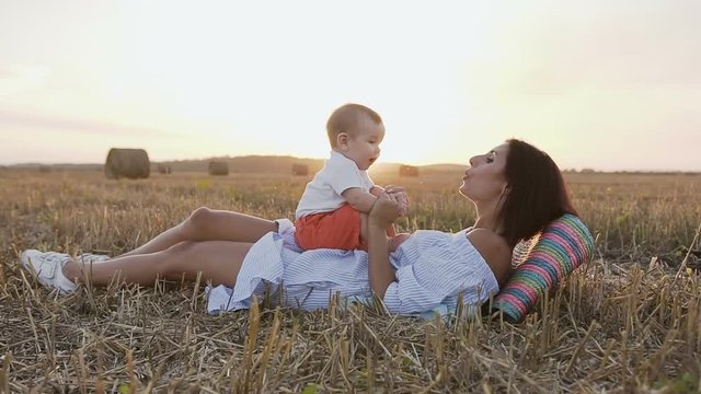 Young woman with her adorable 6 month boy playing outdoors in the field. A young mother holding a small son on her arms lays in the field in the summer in the sunset