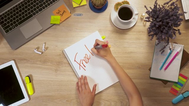 Girl writes on a piece of paper a phrase - Freelance, sitting at work table, first-person view of hands.