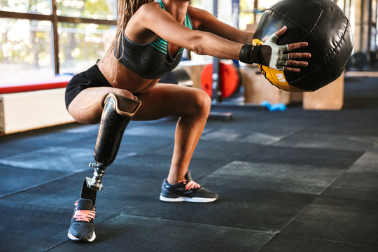 Image of athletic handicapped woman with prosthesis in tracksuit, squatting and holding fitness ball in gym