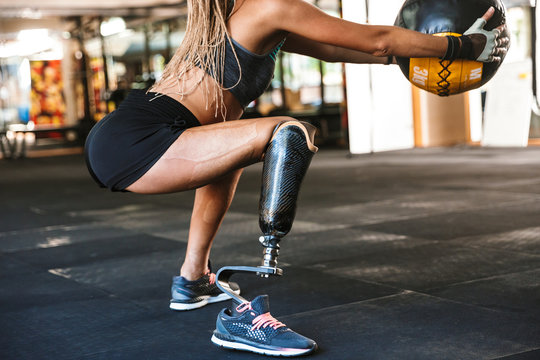 Portrait of beautiful invalid sportswoman with prosthesis in tracksuit, squatting with fitness ball in crossfit gym