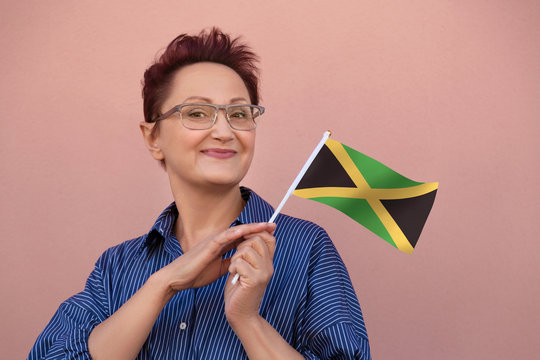 Jamaica Flag. Woman Holding Jamaican Flag. Nice Portrait Of Middle Aged Lady 40 50 Years Old With A National Flag Over Pink Wall On The Street Outdoors.