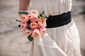 a bridal bouquet of pink, peach, orange austin english rose in the hand of the bride in the background of a stylish wedding dress with black belt . close up