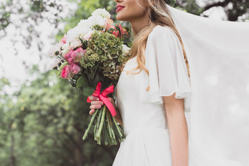 cropped shot of young bride in wedding dress and veil holding beautiful bouquet of flowers