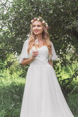 Obraz na płótnie Canvas beautiful smiling young bride in floral wreath and wedding dress holding glass of champagne and looking away outdoors