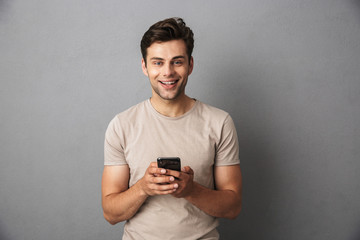 Fototapeta premium Happy young man with short dark hair holding smartphone and chatting, isolated over gray background