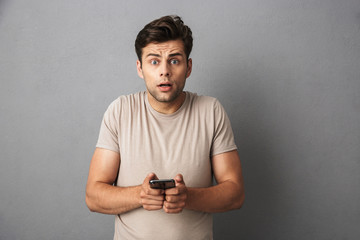 Confused european man with short dark hair holding smartphone and chatting, isolated over gray...