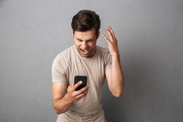Nervous upset guy 20s in t-shirt shouting in anger while looking at smartphone, isolated over gray...